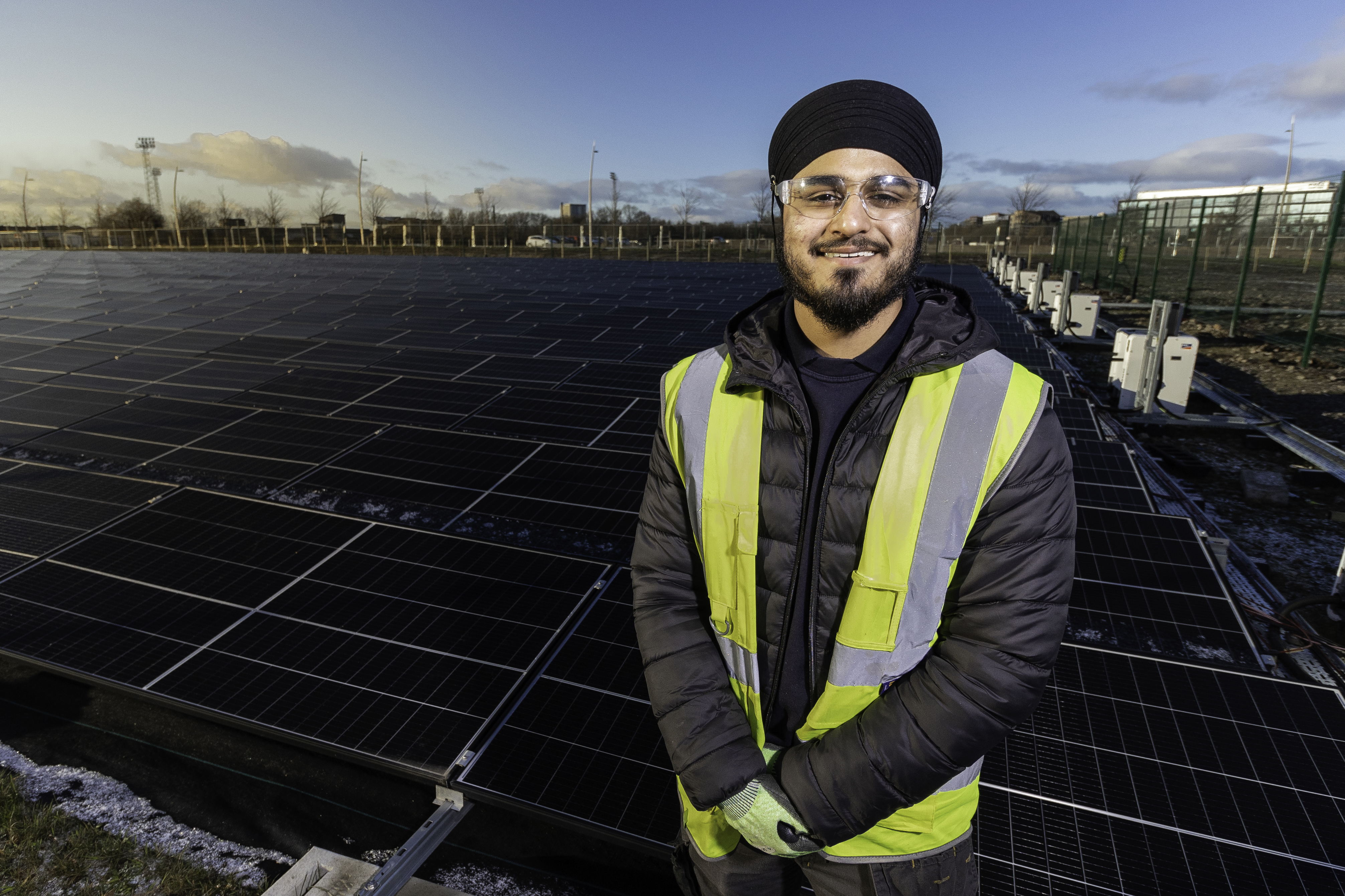 Mandeep smiles to camera, wearing safety goggles, a high vis vest over black work clothes. He stands in front of a field of solar panels