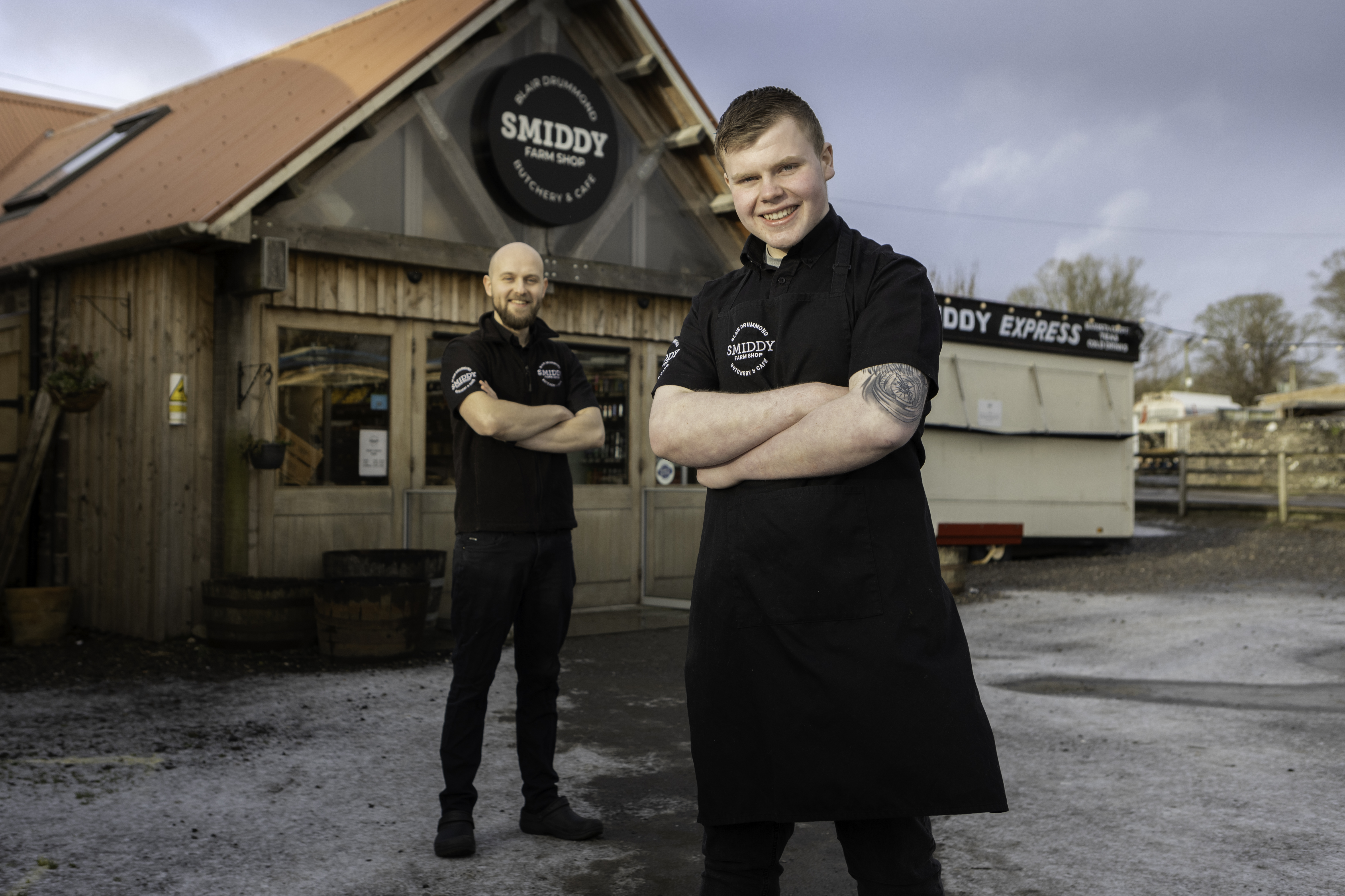 Marc and Fraser stand with arms crossed smiling to camera in front of a wooden clad building that houses the Smiddy Farm Shop where they work.