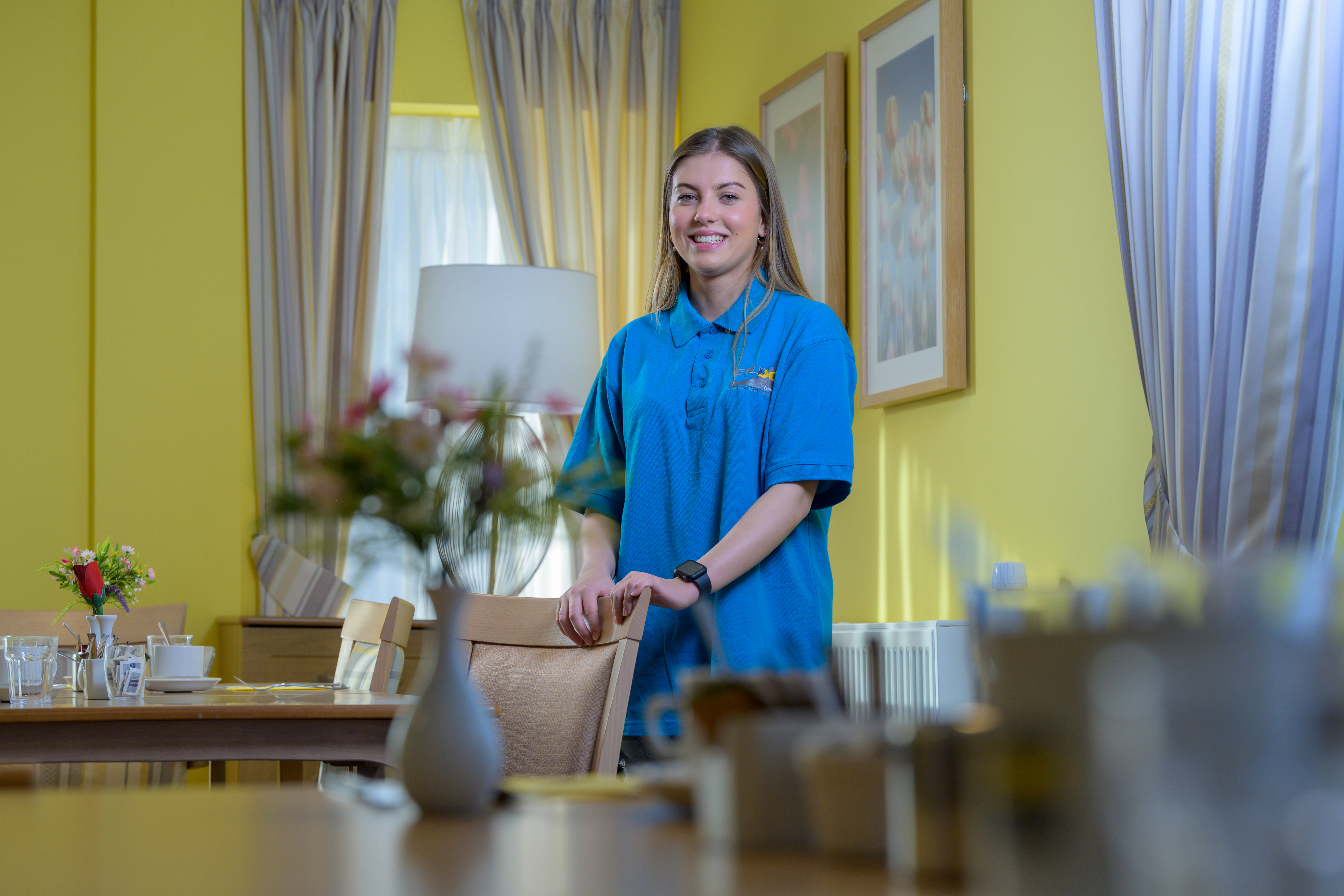 Martyna stands in a sunny dining room, wearing a blue polo shirt. The walls are bright yellow, with vases of flowers on the table.