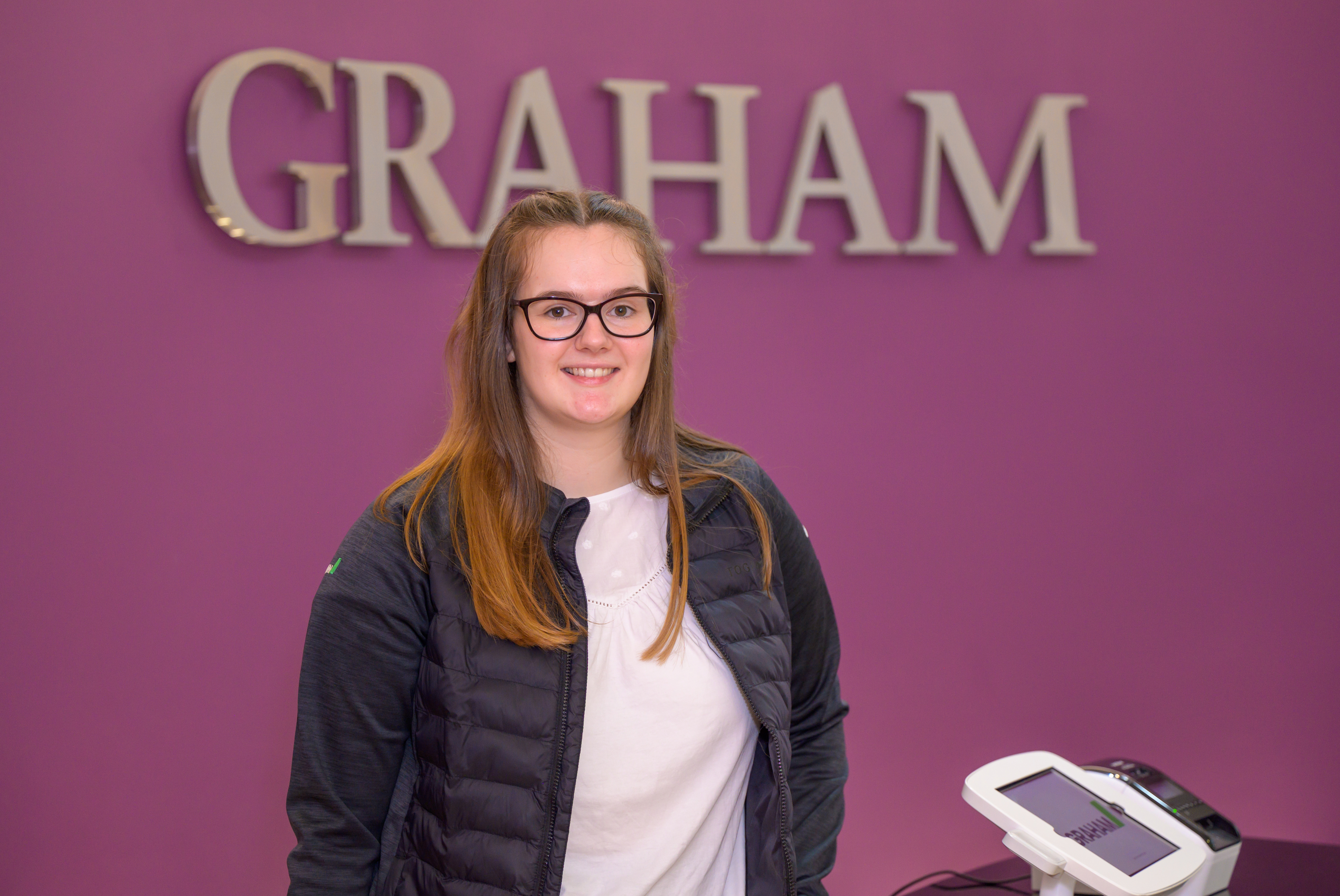 Alexis, with long brown hair and black-rimmed glasses, wears a black jacket and white t-shirt, standing in front of a purple backdrop with her employer GRAHAM's logo in the background. 