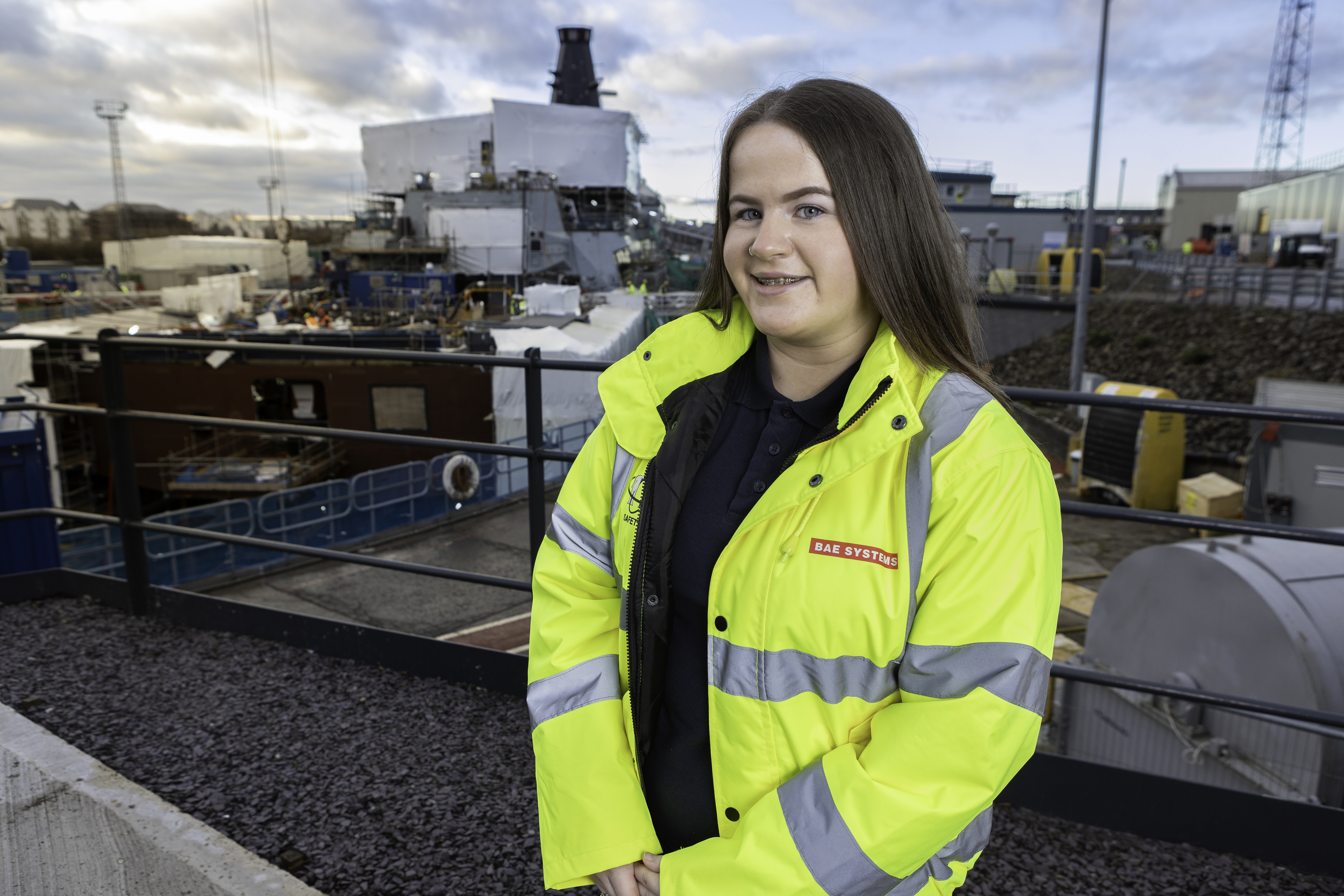 Penny stands smiling at camera, her long brown hair falling over her high vis jacket with a BAE Systems label. In the background is BAE Systems yard. 
