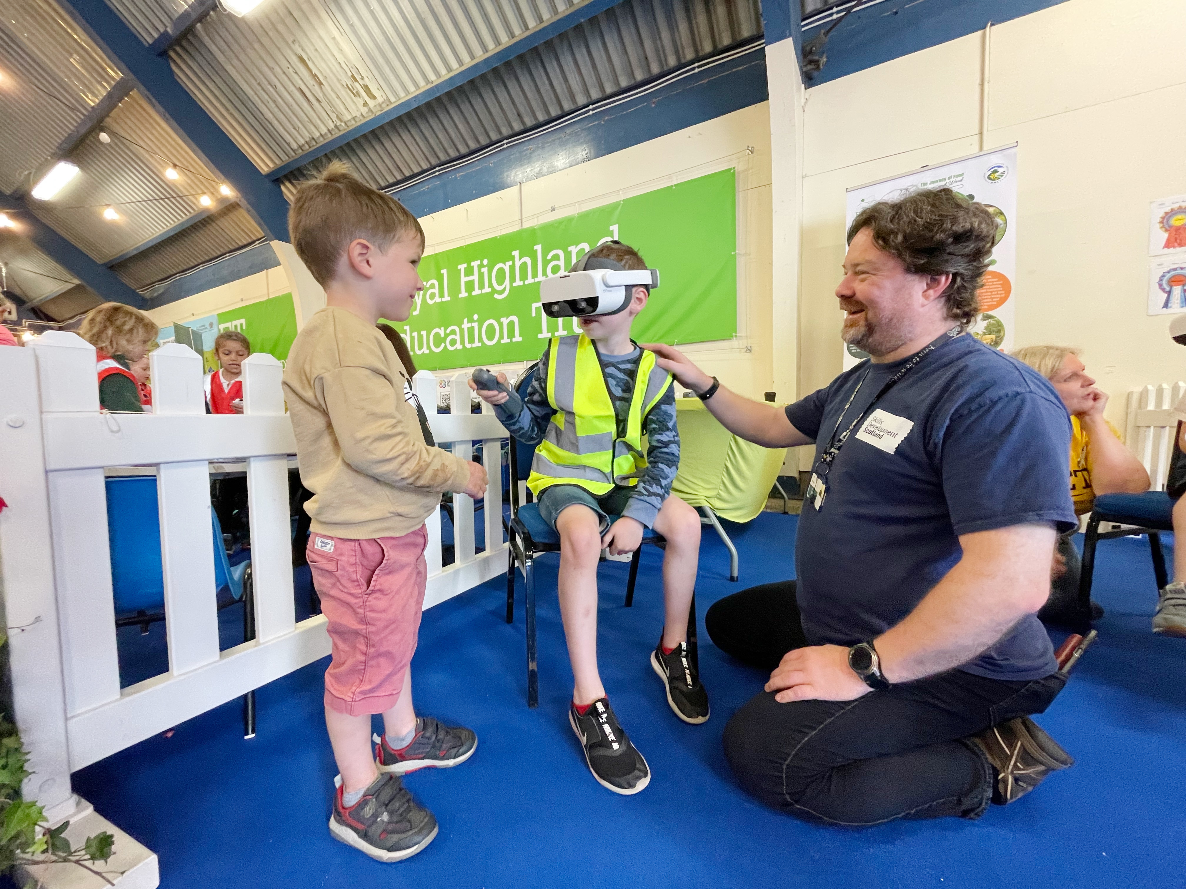 A small boy in a high vis jacket sits in the middle of the picture wearing a VR headset, while his brother and SDS team member look on. 
