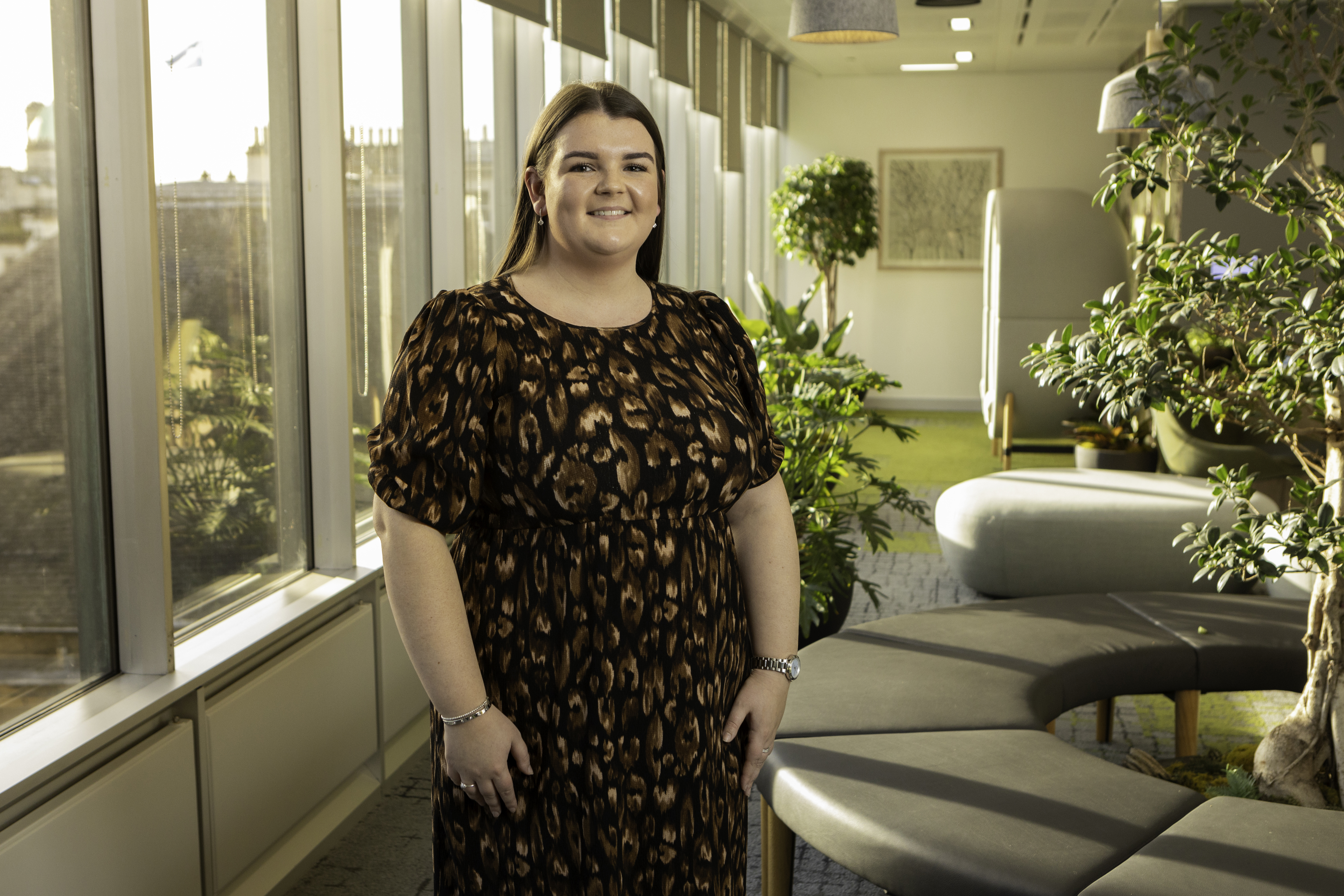 Nicole wears a black and brown dress, standing in a bright office. In the background are relaxed seating areas with plants and trees, and we can see Edinburgh's skyline through windows behind.