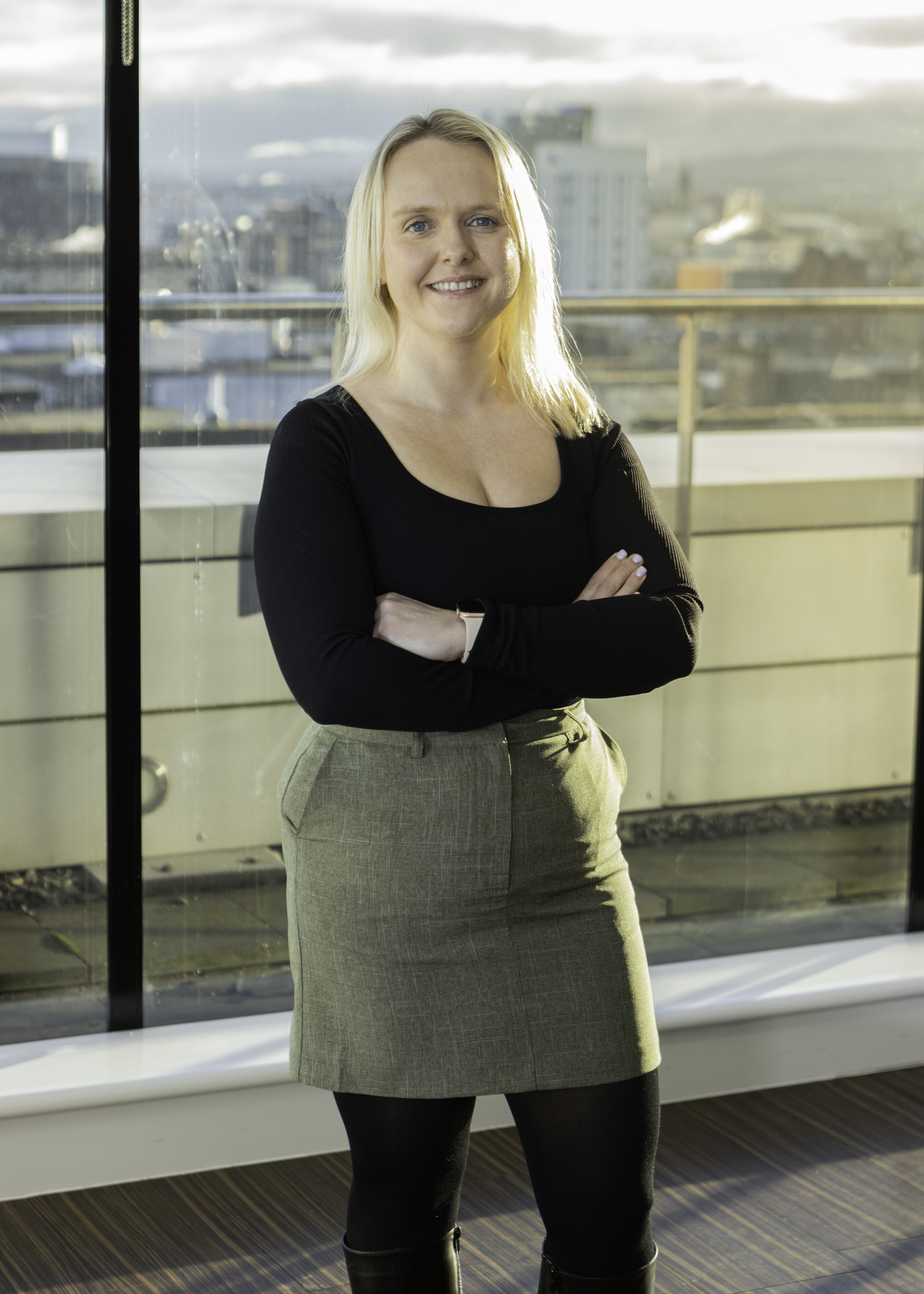 Missy-Ann stands at the window of her office wearing a black top and green skirt. The window behind her shows a city skyline. 