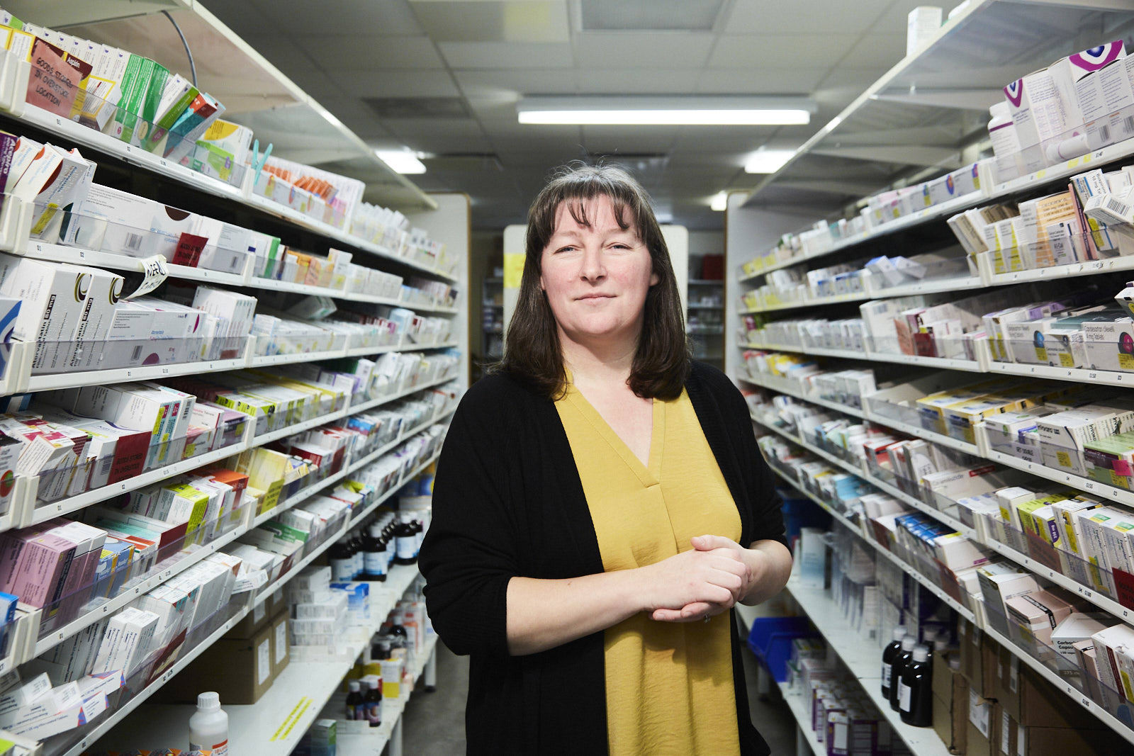 Suzanne wears a pale orange top and black cardigan, she stands between 2 racks of medicine at an NHS pharmacy. 