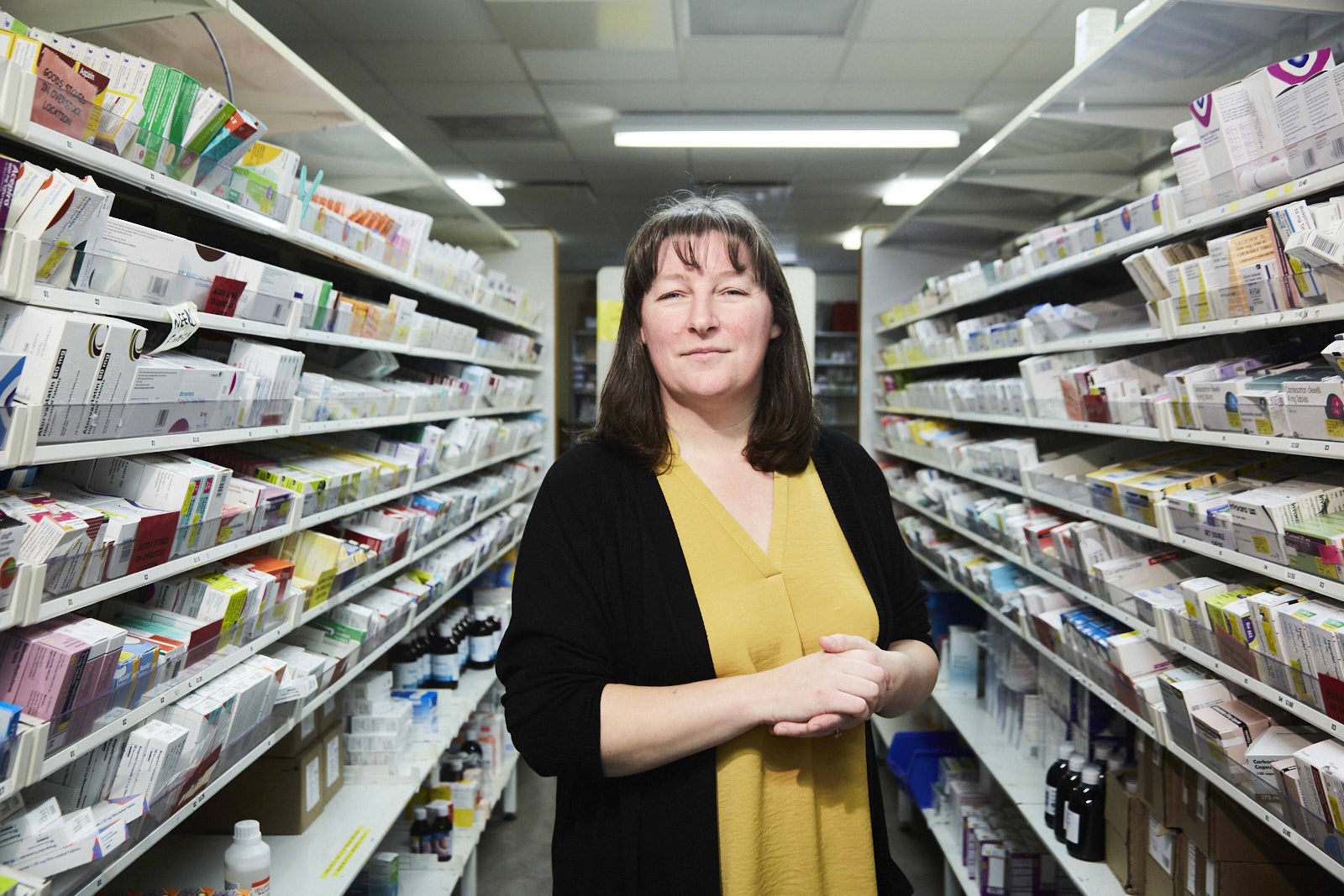 Suzanne wears a pale orange top and black cardigan, she stands between 2 racks of medicine at an NHS pharmacy.